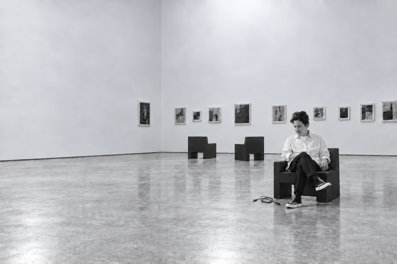 Marcelo Massano sentado em uma cadeira em uma galeria, com duas cadeiras vazias e um cabo no chão, em fotografia em preto e branco.